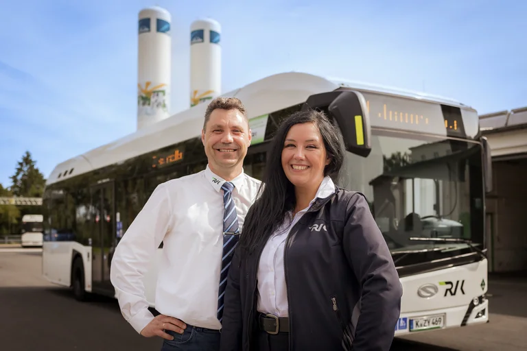 Busfahrerin und Busfahrer stehen gemeinsam posierend und stolz vor einem Bus. Im Hintergrund die zwei Speichertürme für Wasserstoff in Wermelskirchen.