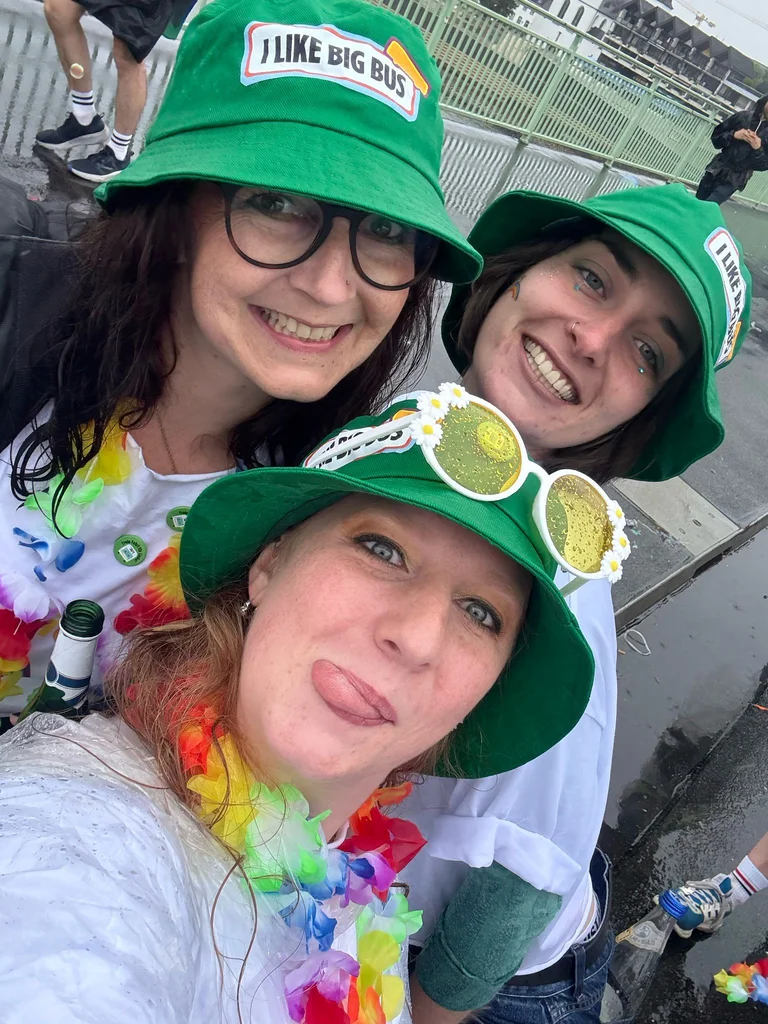 Auf dem Bild sind drei Mitarbeiterinnen zu sehen, die in die Kamera lächeln und grüne Anglerhüte tragen