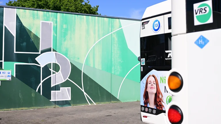 Meckenheimer Wand mit Grafitti in Grüntönen und der Schrift "H2". Davor in der Unschärfe parken zwei Wasserstoffbusse