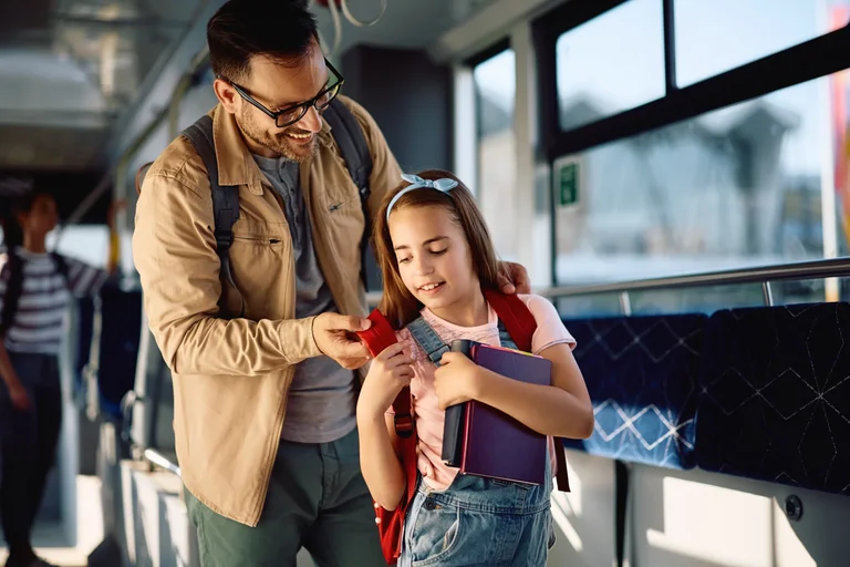 Ein Vater hilft seiner Tochter im Bus stehend den Schulranzen aufzusetzen.