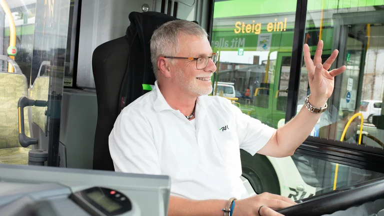 Busfahrer unterwegs. Grüßend hat er die linke Hand beim Fahren des Busses kurz gehoben, um lächelnd seine Kolleginnen und Kollegen zu grüßen. 