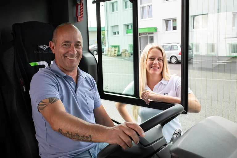 Busfahrer lacht in die Kamera, während er noch auf der Niederlassung unterwegs ist, aber schon Platz in seinem Bus genommen hat. Auf dem Fahrersitz, versteht sich. Eine Kollegin steht am offenen Busfenster und eigentlich halten die beiden noch einen kurzes Gespräch, bevor es raus auf die Straße geht.