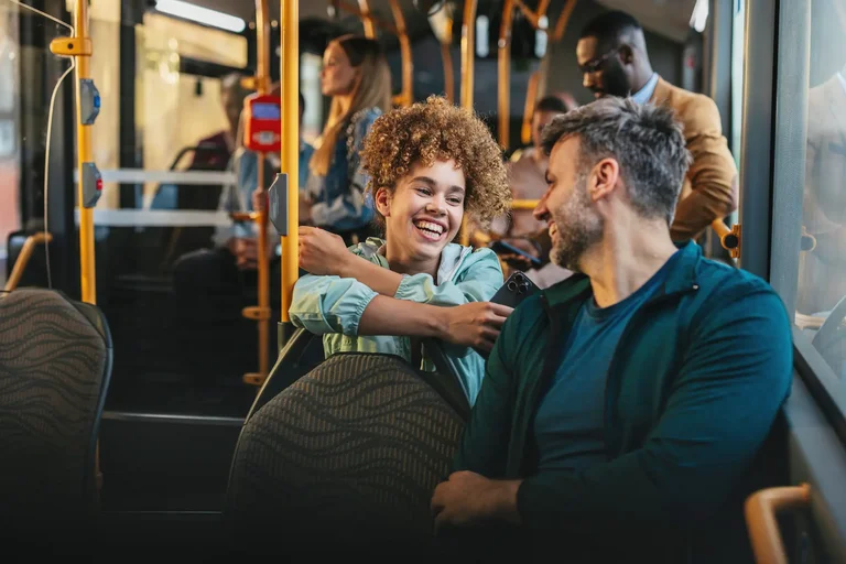 Eine junge Frau und ein junger Mann sitzen hintereinander im Bus, sind einander zugewandt und unterhalten sich lachend.