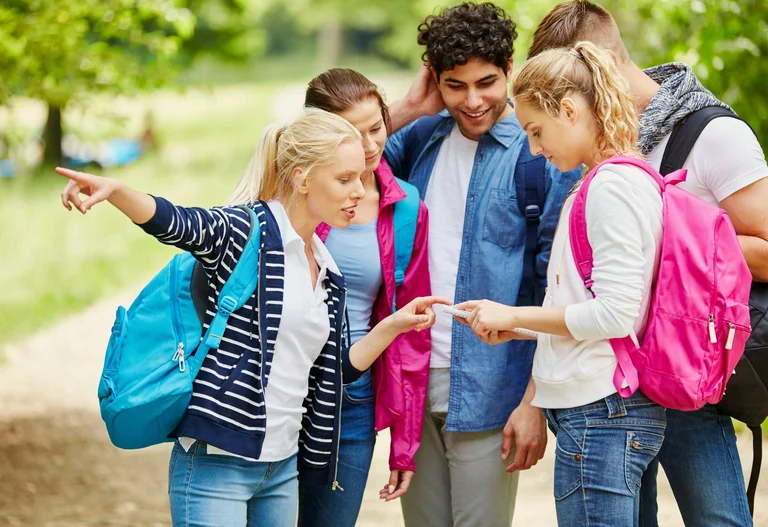 eine junge Menschengruppe schaut gemeinsam auf ein Smartphone, eine junge Frau zeigt mit ihrer Hand in eine Richtung