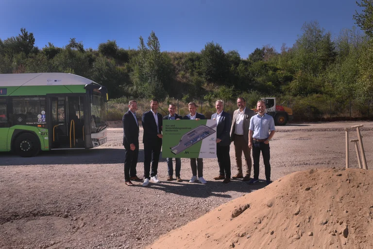 Auf der Baustelle in Mechernich stehen Heiko Iffland (Geschäftsführer Ingenion), Michael Fingel (Bürgermeister Stadt Mechernich), Dr. Marcel Frank (Geschäftsführer RVK), Markus Ramers (Landrat Kreis Euskirchen), Achim Blindert (Allg. Vertreter des Landrates Kreis Euskirchen), Thomas Hambach (Erste Beigeordneter, Stadt Mechernich), Matthias Mohr (Anlagentechnik, RVK) und halten eine Visualisierung der zukünftigen H2-Tankstelle in den Händen. Im Hintergrund steht ein RVK-Bus.