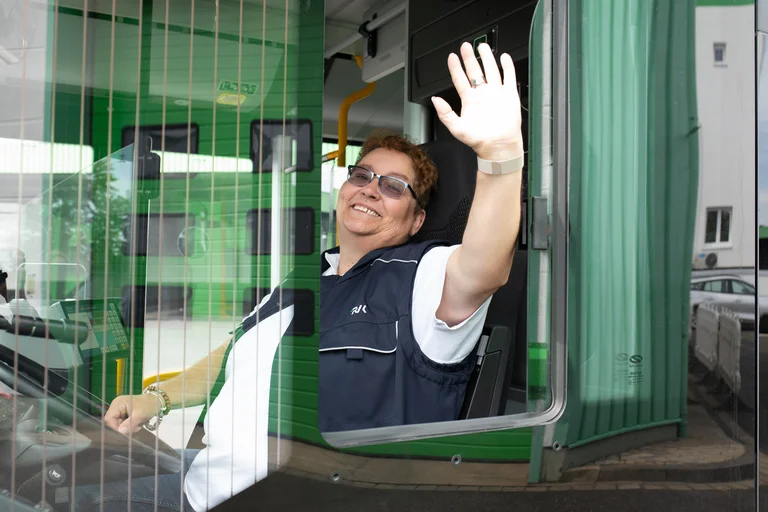 Bis zu beiden Ohren grinsend und mit der linken Hand durchs Fenster winkt die glückliche Busfahrerin während der Fahrt.