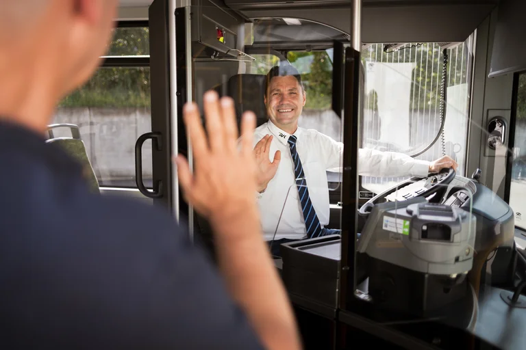 Busfahrer sitzt in der Fahrerkabine und winkt freundlich einem neu einsteigenden Gast.