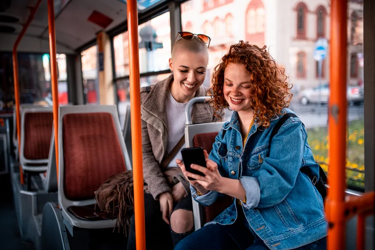 Zwei junge Frauen sitzen hintereinander im Bus und schauen lachend gemeinsam auf ein Smartphone.