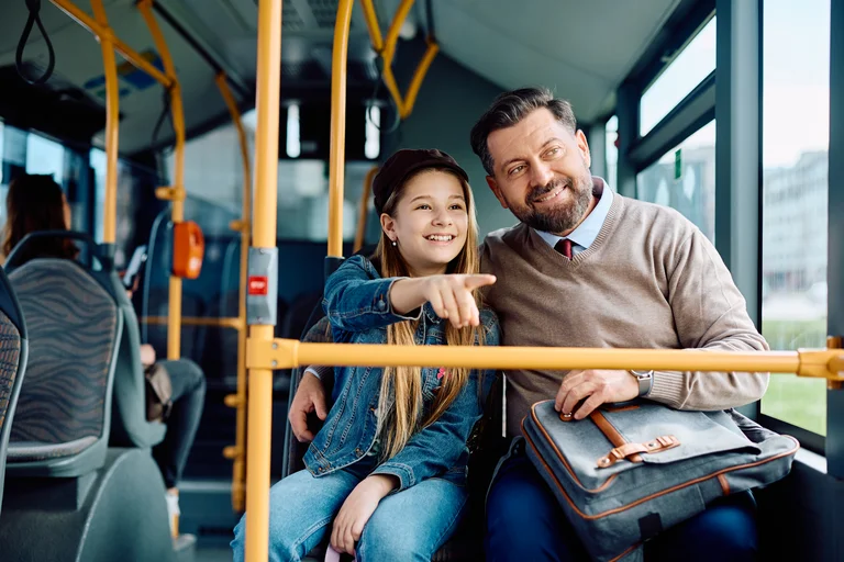Vater und Tochter sitzen nebeneinander am Fenster in einem Bus. Beide schauen gemeinsam aus dem Fenster und die Tochter zeigt auf etwas in der Ferne.