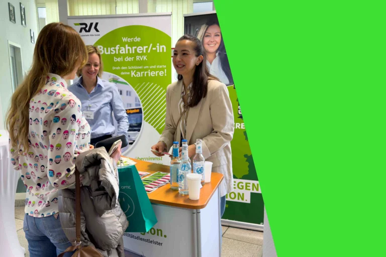 Das Bild zeigt zwei Mitarbeiterinnen der RVK, die lächelnd an einem Infostand eine Frau beraten.