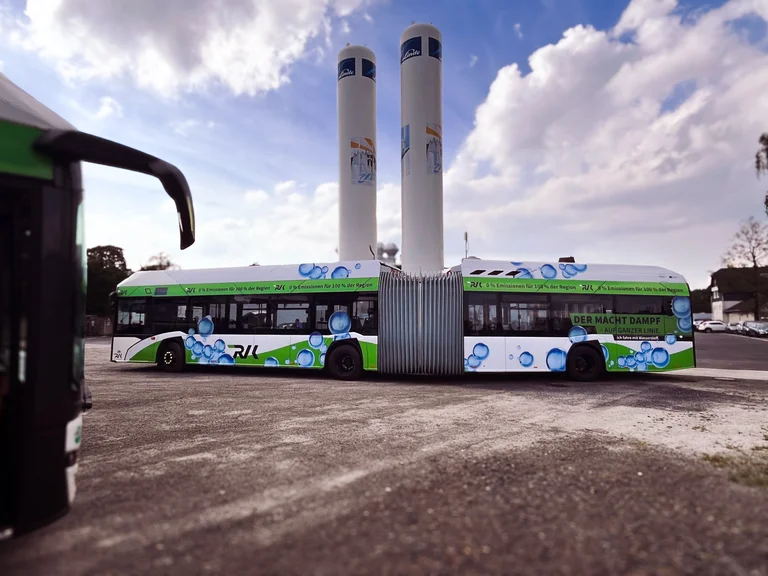Solaris Gelenkomnibus posiert seitlich vor den Wasserstoffspeichertürmen der Wermelskirchener Niederlassung. Der Gom hat eine Folierung aus blauen Wasserstoffblasen und grünen Elementen