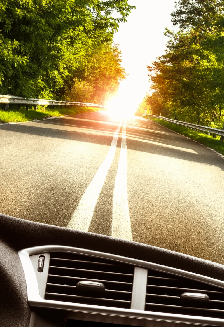 Blick durch die Winschutzscheibe eines Autos. Zusehen ist eine Sraße, die am Hoizont endet mit einer untergehenden Sonne im Hintergrund.