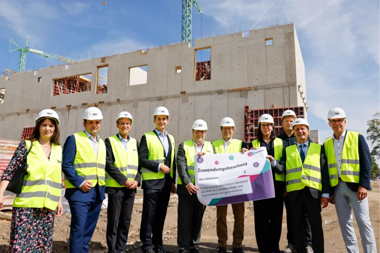 Das Bild zeigt mehrere Menschen mit gelber Warnweste und Bauhelmen, die vor der Baustelle des Grünen Mobilhofs stehen und einen Scheck in die Kamera halten.