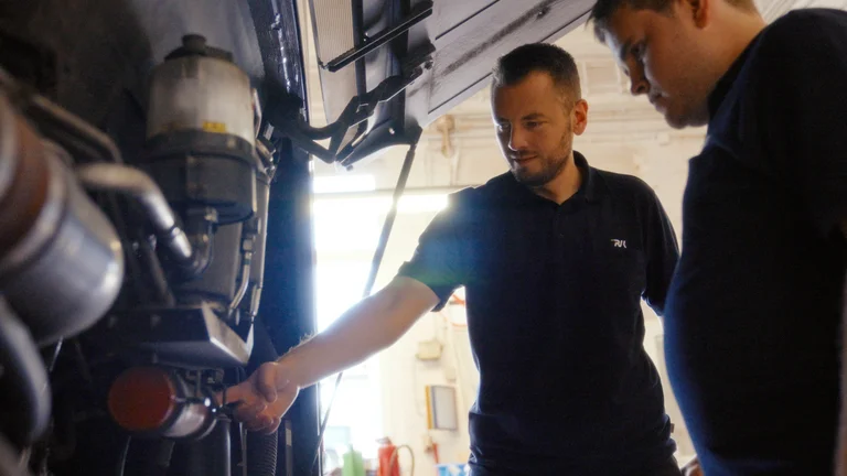 Bus in der Werkstatt und auf der Rückseite geöffnet. Mitarbeitender zeigt einem Auszubildendem etwas.