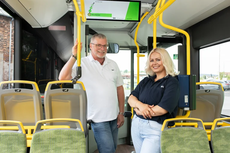 Busfahrerin und Busfahrer posieren in einem RVK Bus, beide schauen lachend in die Kamera.