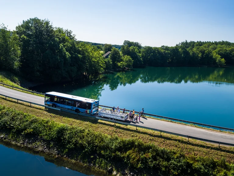 Blick von oben auf eine Talsperre, rechts und links blaues Wasser, drumherum grün. In der Mitte der Straße steht der Bergische Fahrradbus samt Anhänger, auf welchen gerade Fahrräder geladen werden.