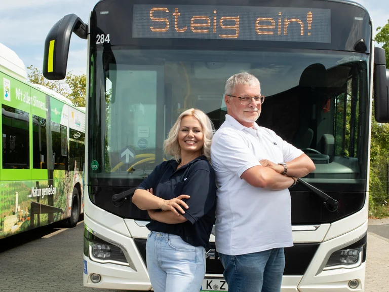 Busfahrerin und Busfahrer stehen Rücken an Rücken lachend vor einem MAN auf der Euskirchener Niederlassung an einem sonnigen Tag. Auf dem Bus prangt die Anzeige "Steig ein!".