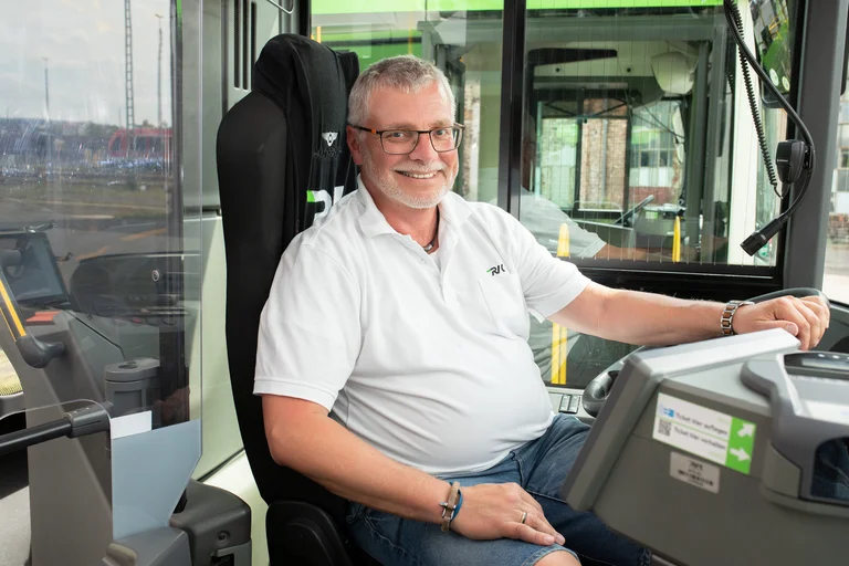 Busfahrer lacht in die Kamera, während er schon eine Hand am Lenkrad hat. Bereit, um gleich in seine Schicht zu starten.