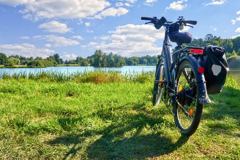 Eine grüne Wiese mit Blick auf einen See. Im Vordergrund steht ein Fahrrad mit Satteltaschen. Der Himmel ist blau mit kleinen Wolken.