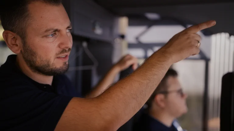 Auszubildender sitzt hinterm Steuer eines Busses, Mitarbeitender zeigt auf etwas in der Ferne