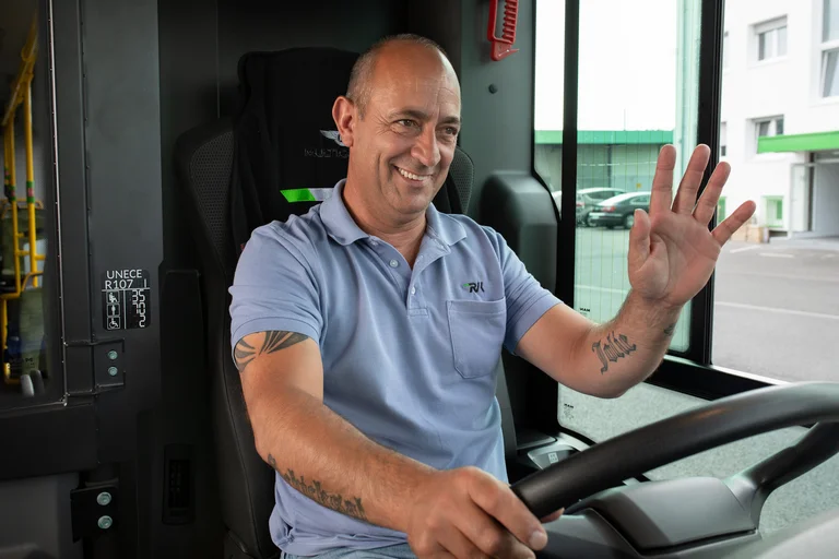 Busfahrer sitzt auf dem Fahrersitz. Er hat die rechte Hand am Steuer, mit der Linken grüßt er lachend vorbeikommende Kolleginnen und Kollegen. 