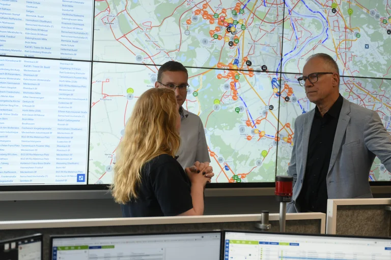 Das Bild zeigt Dr. Marcel Frank und Dr. Norbert Reinkober im Gespräch mit einer Mitarbeiterin vor der großen Videowand der Leitstelle